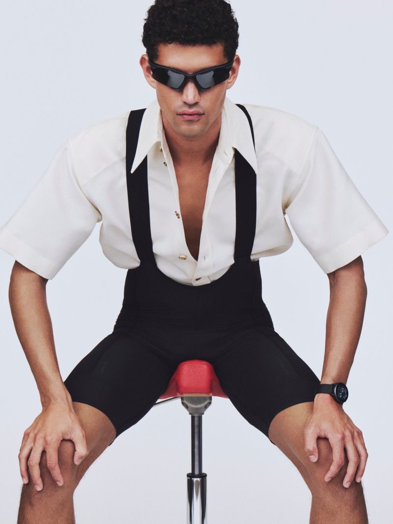Model Bodhi Heeck wearing black sunglasses, an open white shirt, and black cycling bibs seated on a red bike saddle against white.