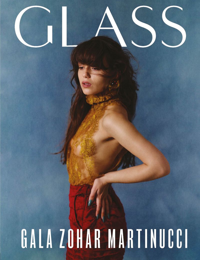 Magazine cover featuring a young woman with bangs, gold lace top, and a red skirt, posing against a muted blue background, with "GLASS" printed above.