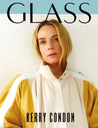Portrait of a woman with blonde hair wearing a white and yellow high-neck garment against a white tiled wall