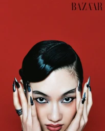 Close-up beauty portrait of Yuemeng Ma with long metallic stiletto nails framing her face against a crimson red background.