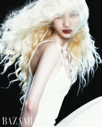Model Yuemeng Ma with voluminous wavy platinum blonde hair and red lips wearing a white tank top against a black background.
