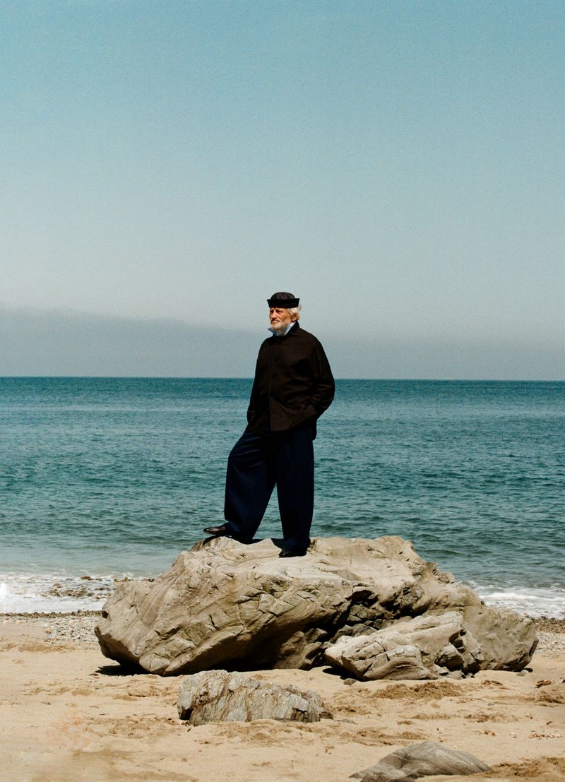 Older man in a dark jacket, wide-leg trousers, and cap standing atop a rock on a sandy beach facing the ocean.