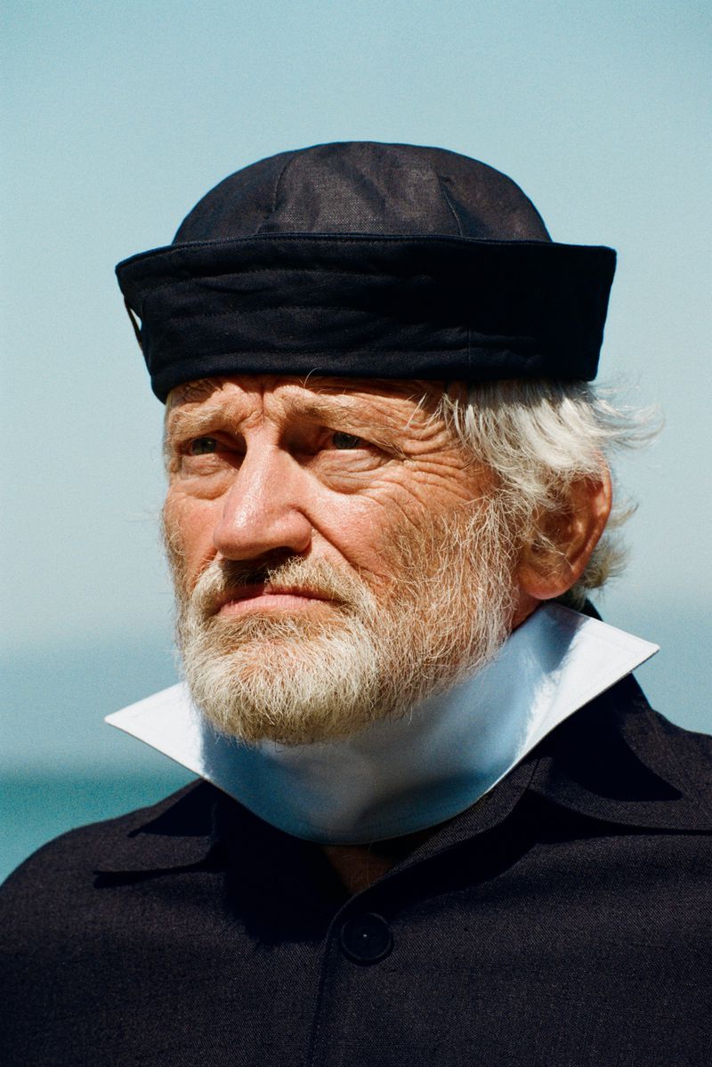 Close-up portrait of an elderly bearded man wearing a black sailor hat and coat with a structured white collar against a blue sky.