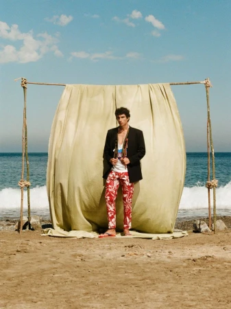 Duncan Yair posing in a black blazer and red floral pants in front of a draped backdrop on a sandy beach.