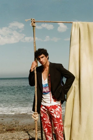 Model Duncan Yair poses on a beach in a black blazer and red printed trousers beside a draped fabric frame.