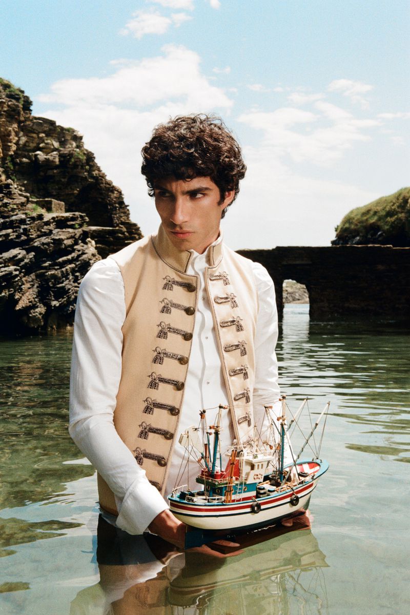 Man in an embroidered beige vest standing in clear sea water holding a miniature fishing boat near rocky cliffs.