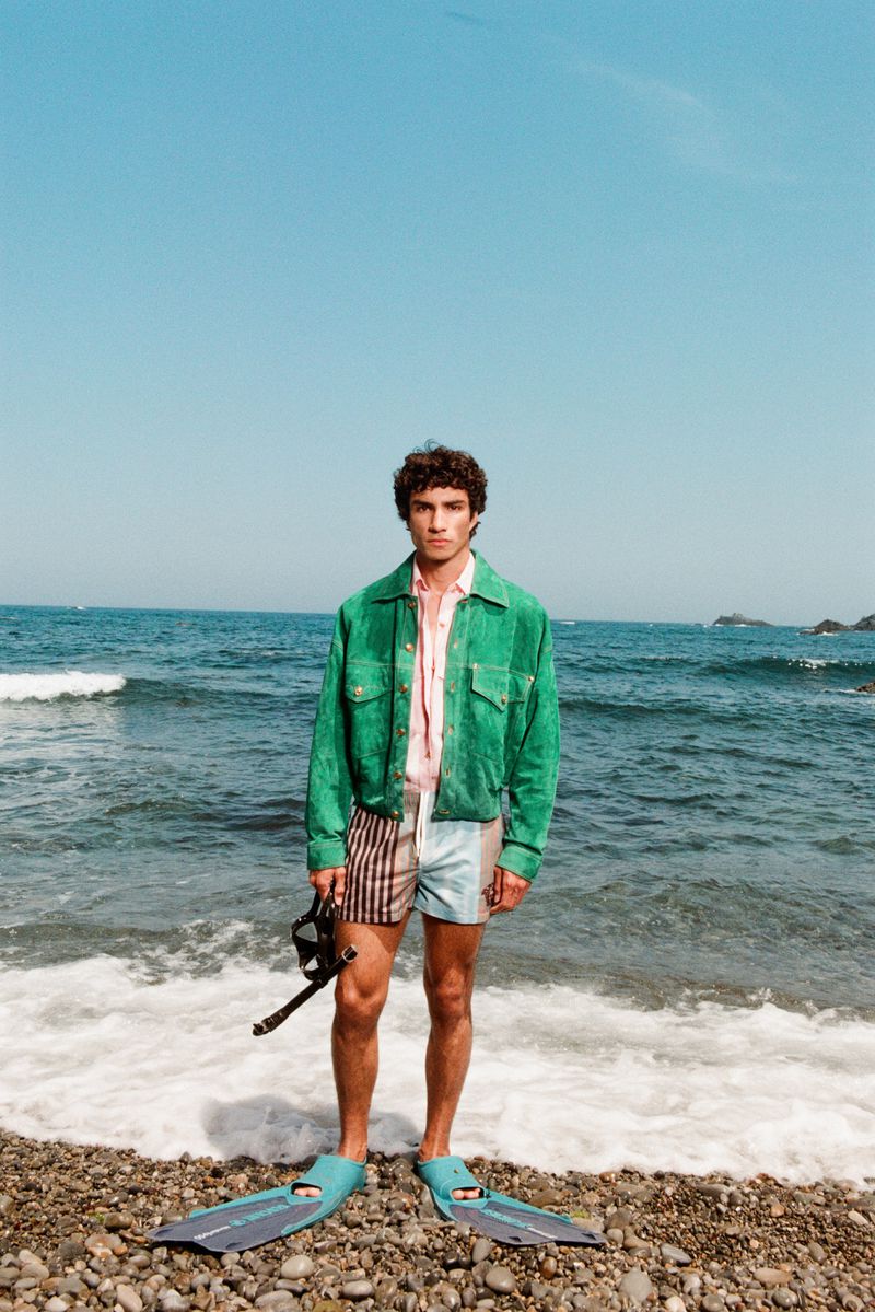 Model Duncan Yair standing on a pebble beach wearing a green jacket, swim trunks, flippers, and holding a snorkel mask.