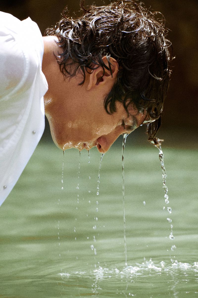 Close-up profile of a man with wet dark hair leaning over green natural water with water streaming from his face.