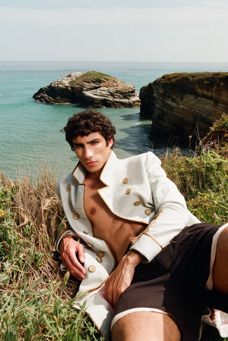 Model wearing a cream military-style jacket reclining on a grassy cliff overlooking the ocean for Esquire Portugal editorial.