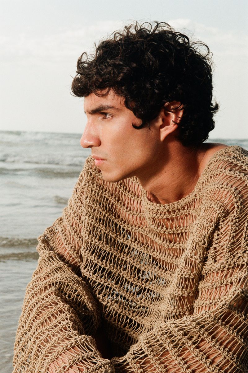 Model Duncan Yair wearing a textured beige open-knit mesh sweater sitting in front of ocean waves on a beach.