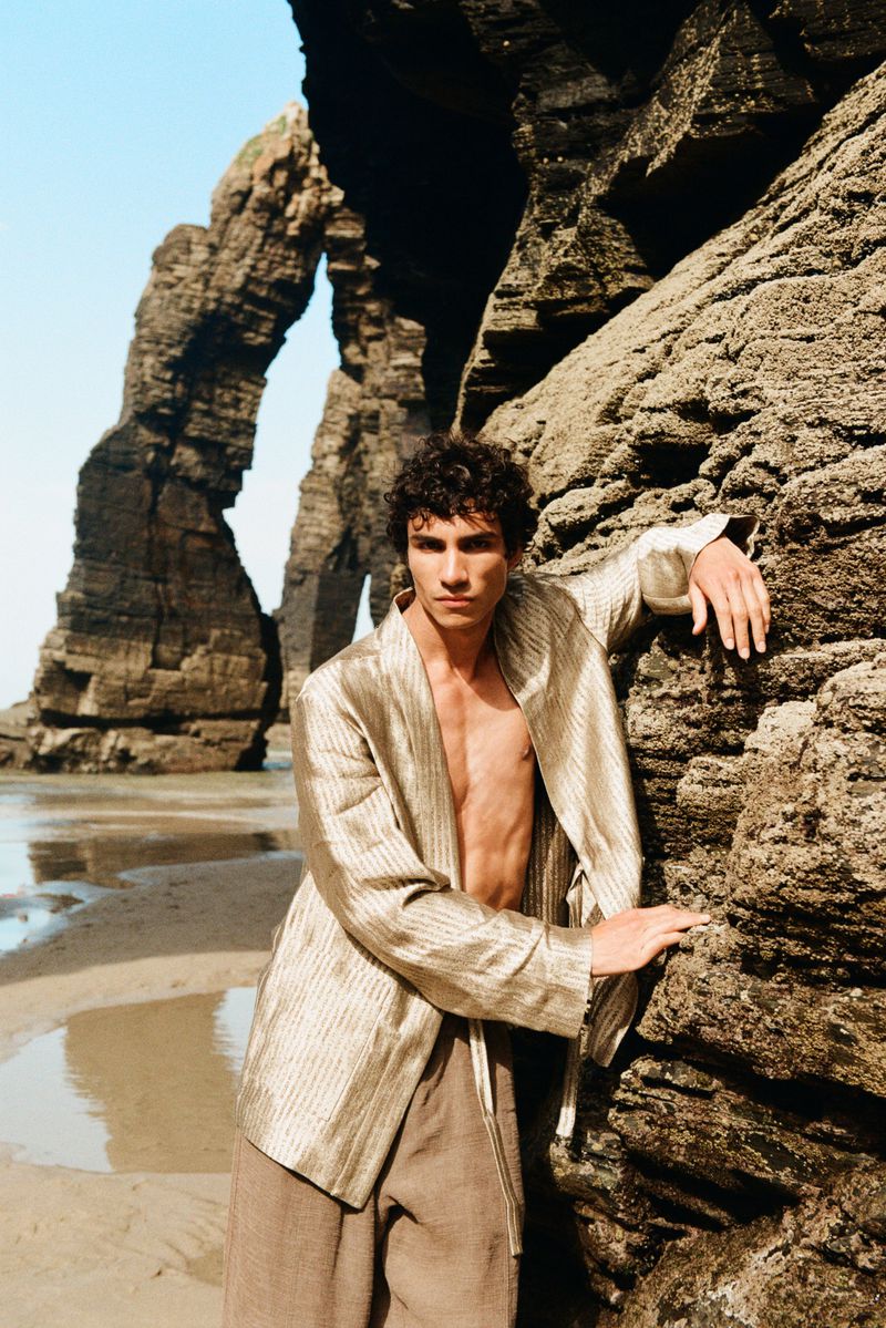 Duncan Yair posing shirtless in a shimmering metallic gold jacket against rugged coastal rock formations on a sandy beach.