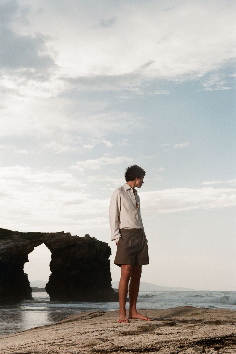 Model Duncan Yair stands barefoot on a rocky coastal cliff wearing an off-white button-down shirt and brown shorts by the sea.
