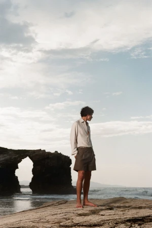 Model Duncan Yair stands barefoot on a rocky coastal cliff wearing an off-white button-down shirt and brown shorts by the sea.