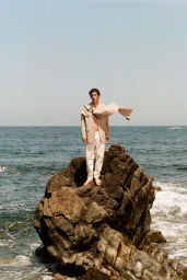 Model Duncan Yair stands barefoot on a jagged coastal rock wearing a beige jacket and white trousers against an open ocean horizon.