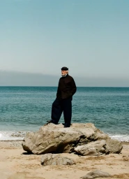 Older man in a dark jacket, wide-leg trousers, and cap standing atop a rock on a sandy beach facing the ocean.