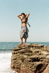 Model Duncan Yair posing atop a rugged coastal rock wearing a sheer mesh crop top and draped grey shorts against an open ocean.