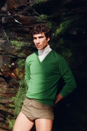 Model Duncan Yair posing outdoors against a mossy rock face in a green V-neck sweater, white turtleneck, and khaki tailored shorts.