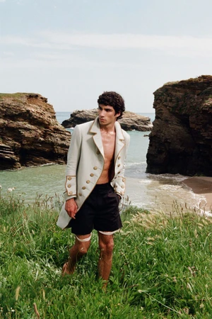 Model Duncan Yair wears an unbuttoned cream military frock coat and black shorts on a grassy cliff above a rocky cove.