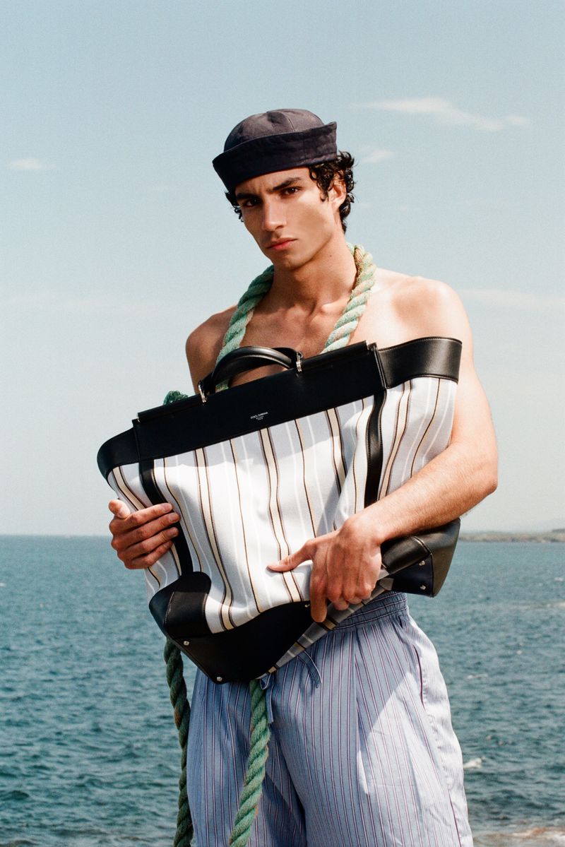 Duncan Yair shirtless outdoors wearing a navy sailor hat and holding an oversized striped tote bag against the sea.