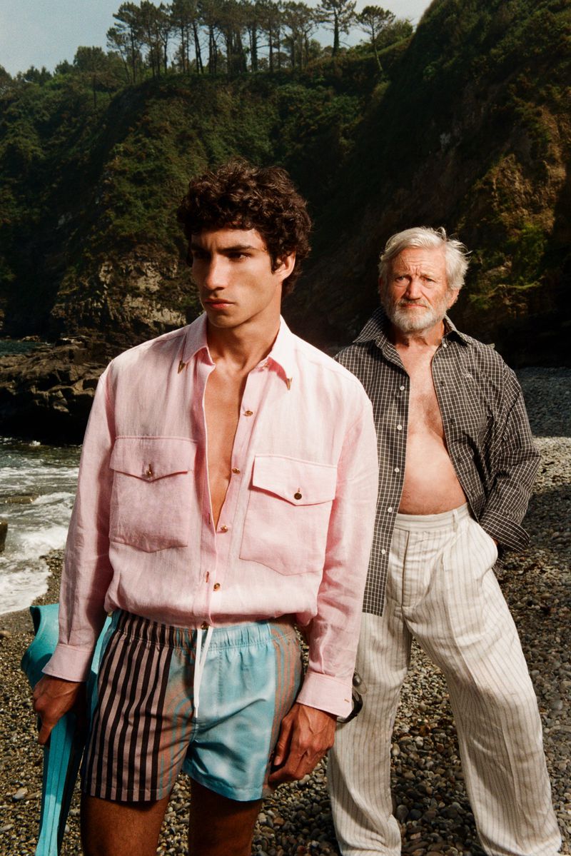Two men posing on a rocky shoreline in resort wear, featuring a young man in a pink linen shirt and patterned swim shorts.