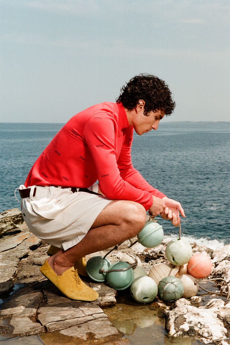 Duncan Yair crouches on a rocky shoreline holding fishing buoys, wearing a bright red long-sleeve top and white shorts overlooking the ocean.