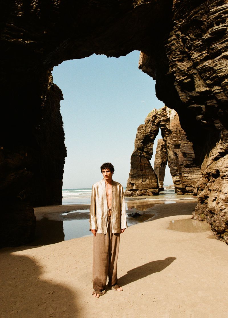 Model Duncan Yair in a loose open shirt and wide linen trousers standing beneath dramatic coastal rock arches on a sandy beach.
