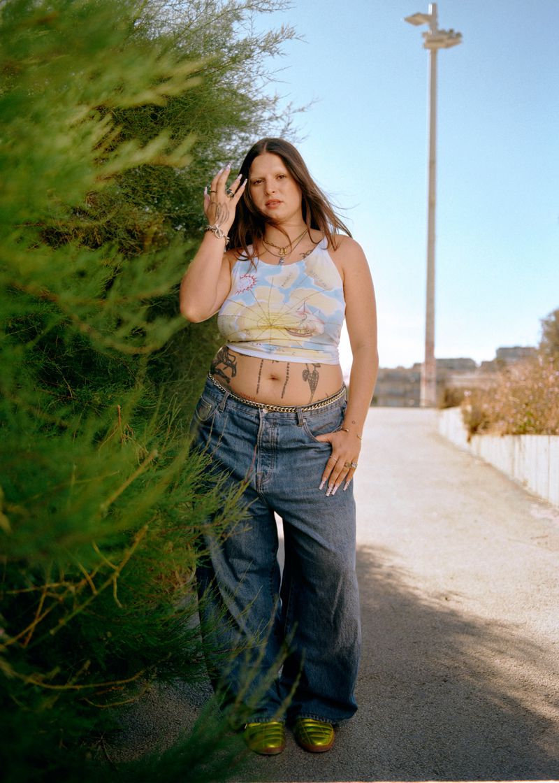 A woman in a floral crop top and baggy blue denim jeans stands beside green foliage on a paved outdoor path.