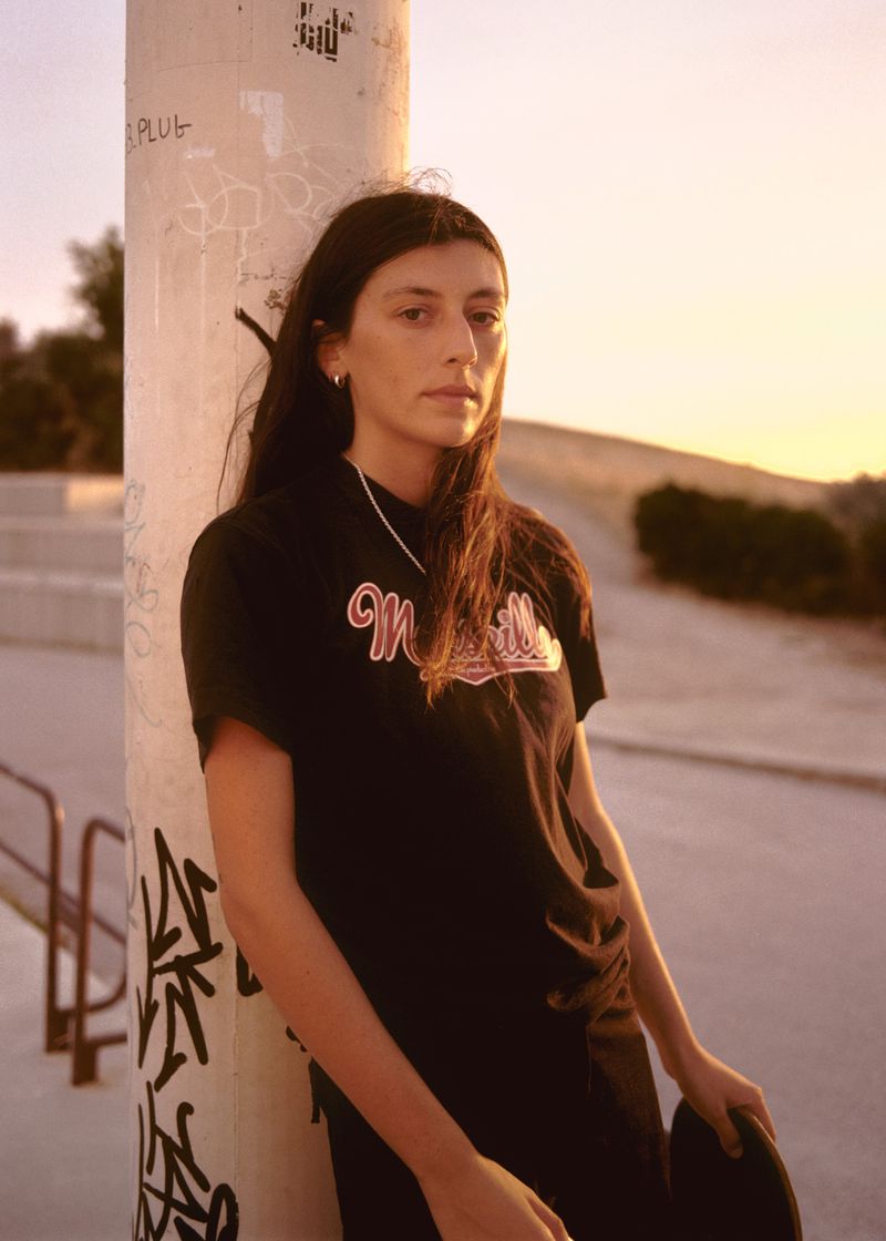 A woman in a black graphic t-shirt leaning against a graffiti-covered pole holding a skateboard at golden hour.