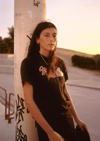 A woman in a black graphic t-shirt leaning against a graffiti-covered pole holding a skateboard at golden hour.