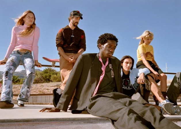 Group of fashionable young adults gathered at an outdoor skate bowl under a clear blue sky, featuring a man in a suit with a pink tie.
