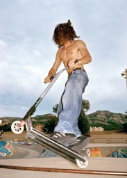 Shirtless person in baggy blue jeans performing a trick on a silver stunt scooter at an outdoor skatepark bowl.