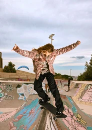A model skateboarding across a graffiti-covered concrete ramp wearing a pink patterned jacket and black trousers at a skatepark.