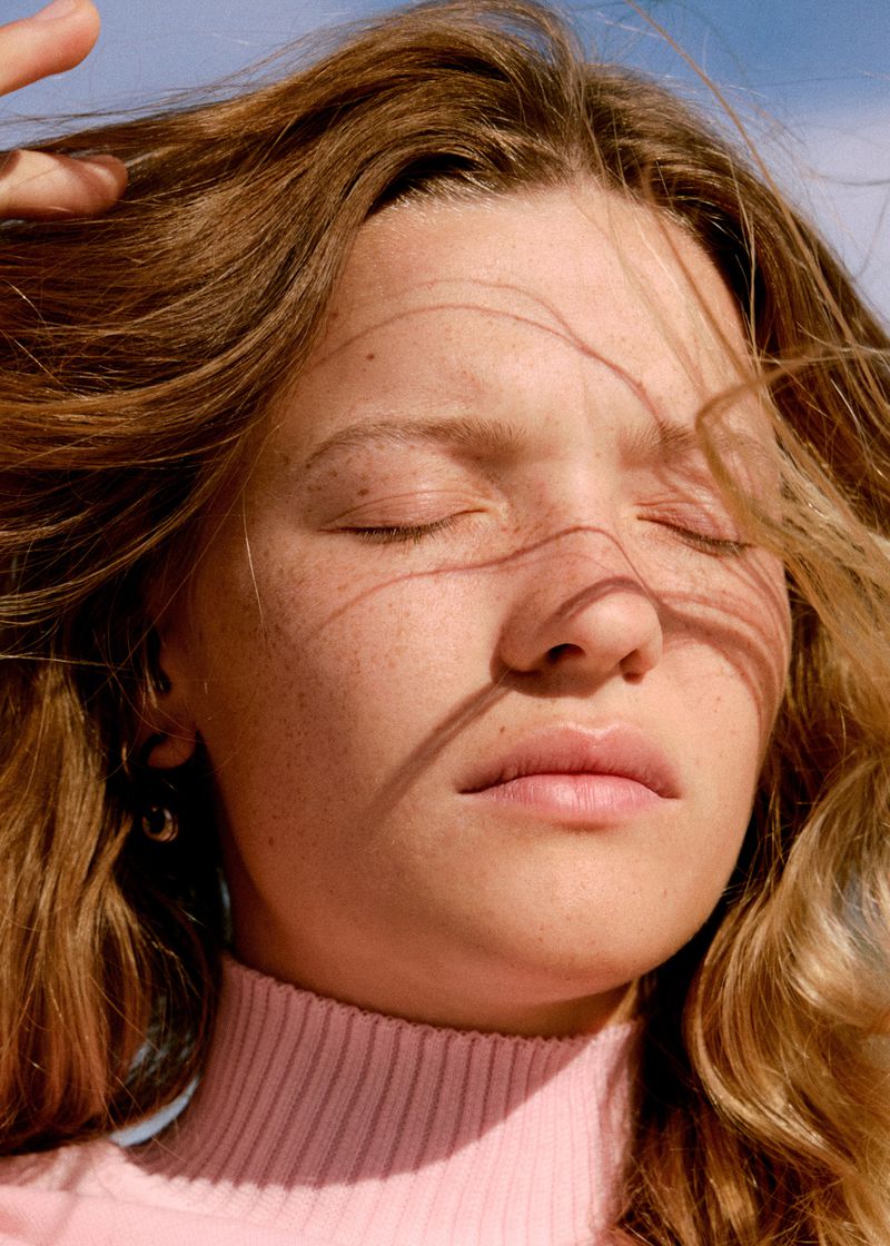 Close-up portrait of a woman with closed eyes, windblown brown hair, and freckles, wearing a pink ribbed turtleneck.