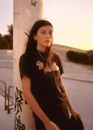 A woman in a black graphic t-shirt leaning against a graffiti-covered pole holding a skateboard at golden hour.