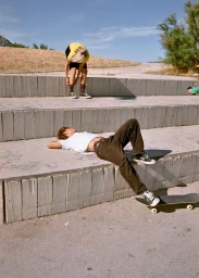 Young man lying back on outdoor concrete steps near a skateboard while another person stands on the tier above in direct sunlight.