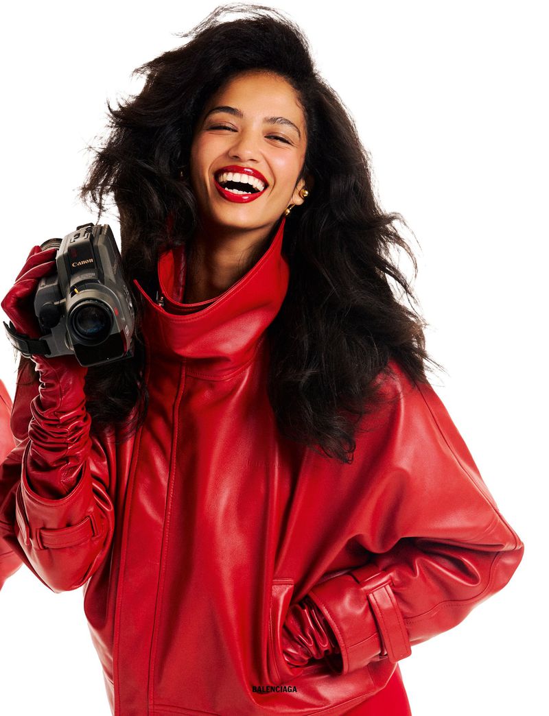 Model Jessie Aina laughs while holding a vintage camcorder, wearing an oversized red leather jacket and matching gloves against white.