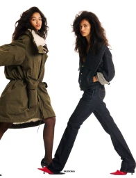 Two fashion models pose against a white background, one in an olive green parka and one in a dark denim set with red pointed heels.