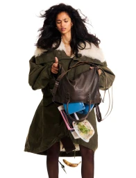Model Jessie Aina in an oversized green shearling parka looking into an overflowing brown leather handbag against a white background.