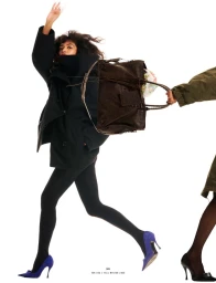 Model Jessie Aina in a black coat, tights, and purple heels running with a brown Balenciaga bag on a white seamless background.