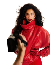 Model Jessie Aina wearing an oversized red leather jacket and matching gloves against a white studio background, seen past a camcorder.