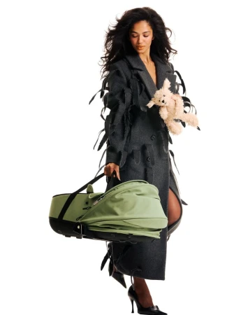 Model Jessie Aina walking in a textured charcoal coat carrying a green bassinet and plush toy against a white background.