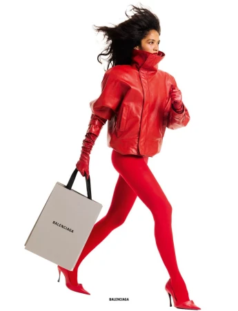 Model Jessie Aina in an all-red leather jacket, gloves, tights, and heels carrying a grey Balenciaga shopping bag against a white backdrop.