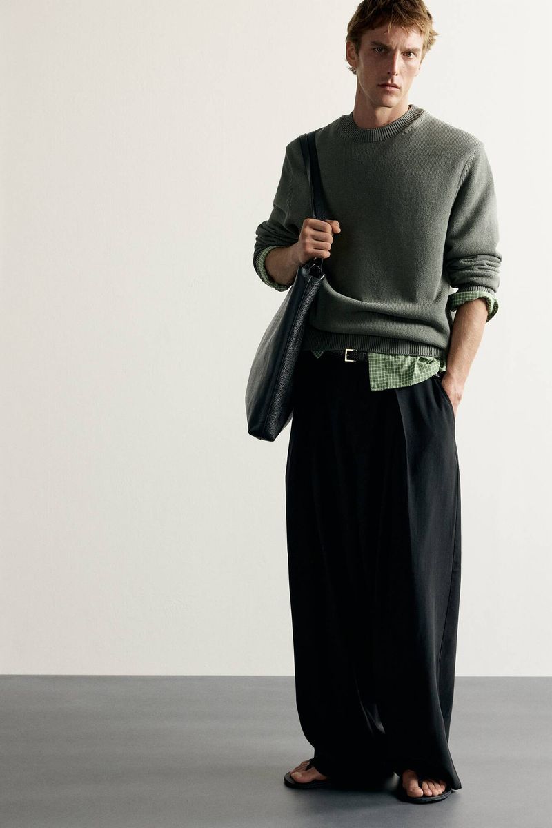 Model posing in a green crewneck sweater, black wide-leg trousers, and sandals with a black shoulder bag against a neutral backdrop.