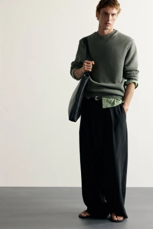 Model posing in a green crewneck sweater, black wide-leg trousers, and sandals with a black shoulder bag against a neutral backdrop.