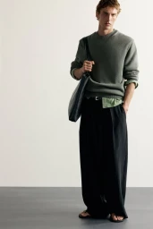 Model posing in a green crewneck sweater, black wide-leg trousers, and sandals with a black shoulder bag against a neutral backdrop.