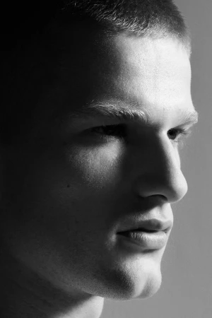 Black and white close-up side profile portrait of a young man with a short buzz cut against a plain grey studio backdrop.