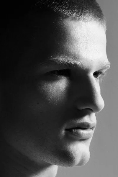 Black and white close-up side profile portrait of a young man with a short buzz cut against a plain grey studio backdrop.