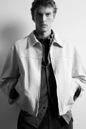 Monochrome portrait of a man wearing a light zip-up jacket over an unbuttoned collared shirt against a plain studio background.