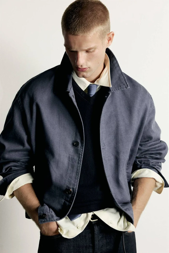Male model with a buzz cut wearing a layered blue jacket, navy sweater, and collared shirt with a tie against a white backdrop.