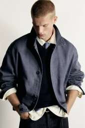 Male model with a buzz cut wearing a layered blue jacket, navy sweater, and collared shirt with a tie against a white backdrop.
